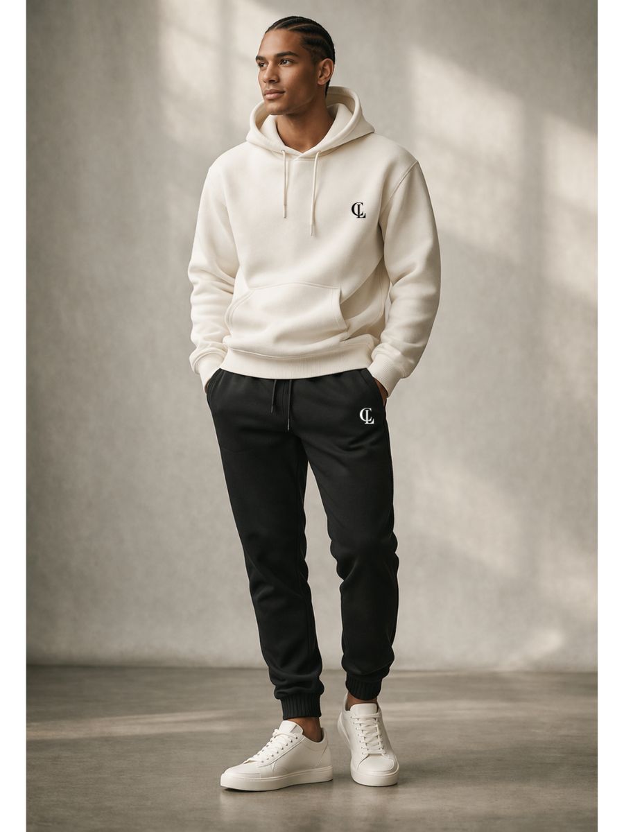 Man wearing a white hoodie and black pants with a logo, standing against a neutral background